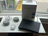 Photo showing the lot items arranged on a white surface by a window: two gray Google Home Mini speakers with cords, white Apple Magic Mouse, black Samsung Blu-ray player, and boxed white b-MOLA air filter.