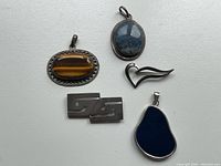 Photo showing five vintage sterling silver jewelry pieces including pendants with tiger's eye, blue stones, an abstract heart-shaped brooch, and a geometric rectangular brooch.