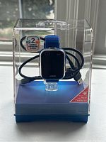Front view of VTech smart watch in clear plastic box featuring blue strap and USB cable, showing dual cameras and screen details.