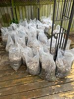 Multiple transparent plastic bags filled with river rock and some soil, placed on a wooden deck outside near a wooden fence.