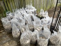Multiple full clear plastic bags filled with river rock and some soil, arranged in rows on a wooden deck with a wooden fence background.