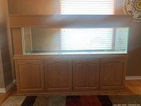 Front view of the large fish aquarium and wooden cabinet stand against a wall with a window behind, showing overall condition and structure.
