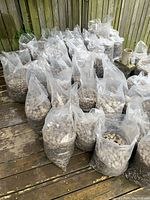 Multiple large clear plastic bags filled with light-colored river rocks, some soil present, arranged on wooden decking near a fence.