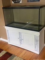 Front view of a large rectangular glass aquarium on a white wooden cabinet stand with two doors. Aquarium has black trim on edges and transparent glass.