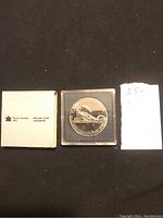 Photo of 1986 Canadian silver dollar coin in plastic case with Royal Canadian Mint paper packaging to the left and a small white paper with handwritten numbers to the right.