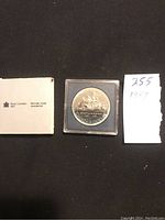 Photo showing the 1987 silver dollar coin in a plastic case with accompanying Royal Canadian Mint box and handwritten note showing '255 1987'.