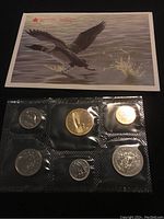 Seven Canadian coins in sealed plastic holder, arranged in two rows with one row containing four coins, the other three. Above holder is a picture of two geese flying over water.