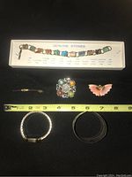 Photo of all six items arranged on black surface with measuring tape showing size. Includes charm bracelet on labeled card, butterfly pin, decorative pen, gold-toned pin, silver-toned bracelet, Timex watch with decorative band.