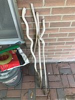 Four wooden trellis sticks leaning against a brick wall next to a small galvanized steel garbage pail with lid and a bag of soil or fertilizer.