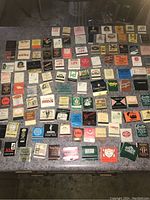 Photo showing a large assortment of over 100 vintage matchboxes and matchbooks laid out on a surface in rows. Various brands and designs visible.
