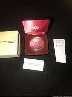 1980 silver dollar coin in protective capsule inside red velvet-lined display box with maple leaf emblem on the lid, alongside original paperwork and handwritten note.