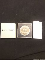 1985 Canadian silver dollar coin in square plastic case, with Royal Canadian Mint booklet and handwritten note beside it