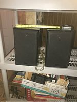 Two KEF black bookshelf speakers side by side with a measuring tape on top showing width approximately 7 inches