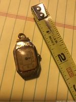 Vintage Waltham pendant watch case placed next to a measuring tape showing watch length slightly less than 2 inches. The crystal face is yellowed and scratched with visible damage.