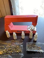 BeDazzler tool shown with five white plastic attachments and two bags containing metal staples and gems on a dark surface.