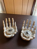 Two ceramic skeleton hand candle holders shown upright with spiderweb candles placed in palms.