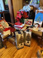 Photo showing chairs with assorted Christmas table linens, boxed fiber optic trees, faux poinsettias in red and silver, ornaments, and some holiday accessories like a Santa hat and stocking.