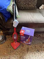 Red Dirt Devil Versa Steam steam mop standing on carpet next to original purple/red box with product branding