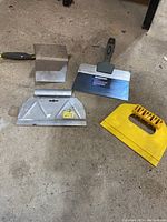 Overview photo showing four drywall and painting tools: a metal corner tool with black handle, a large metal drywall spreader with black handle, a large yellow plastic drywall spreader, and a metal adhesive spreader with wide blade and metal handle.