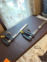 Two Mastercraft 4x12 inch F-Clamps placed on a dark surface next to a window, showing their full length and yellow-handled tightening screws.