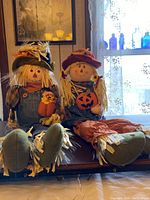 Photo of two seated scarecrow dolls dressed in autumn attire positioned side by side on a table.
