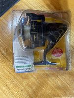 Black and gold Shakespeare Duo-Cast fishing reel in unopened clear plastic packaging with yellow background card.