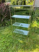 Front view of metal shelving unit with 3 glass shelves visible, set outdoors on grass with green shrubs in background.