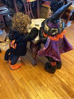 Photo showing two Halloween child decorations standing, one dressed as a witch in purple outfit and hat, the other as a black cat with 'BOO!' on back.