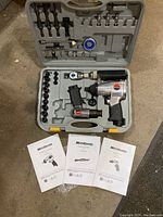 Opened tool kit case displaying two air powered pneumatic impact wrenches, an air chisel and organized sockets and bits, set in grey plastic carrying case, three instruction manuals arranged in front.