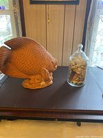 Side view of the terra cotta fish statue and the glass jar filled with shells on a dark surface against a wood-paneled wall with curtains.