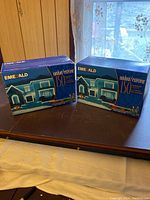Two unopened blue boxes of Emerald 150 outdoor ribbon lights on a table near a window with sheer curtains.