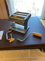 Stainless steel deluxe pasta maker on table with separate wooden handle crank and clamp for securing to surface.