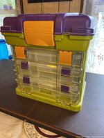 Front view of the plastic organizer showing large top container with purple lid and multiple transparent sliding drawers with purple and orange latches on the front.