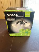 Front view of the NOMA Motion Sensor Security Light box showing the product image, branding, and features such as adjustable detection range and outdoor security use.