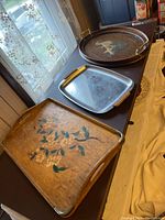 Photo showing three serving trays: wood tray with floral hand-painted design and metal corners, metal tray with wood handles, and wood tray with glass covered picture and metal handles, arranged on table near window.