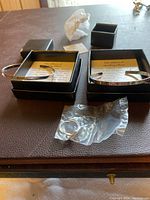 Two sterling silver cuff bracelets engraved with words and two small black presentation boxes with information cards inside, placed on a dark surface.