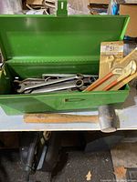 Open green metal toolbox displaying assorted metal wrenches, a pipe wrench, a wooden-handled hammer, and a rubber mallet on a table surface.