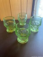 Four green uranium glass water cups with geometric faceted design on a dark surface, shown in natural light.