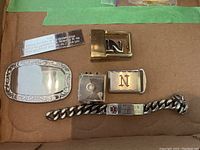 Lot items arranged on cardboard with two gold-tone buckles, one ornate silver buckle, small clip, padlock charm, hospital chain bracelet, and engraved plaque.