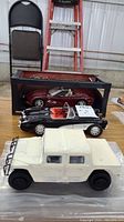 Three model cars on table: white resin Hummer in front, black 1957 Corvette convertible beside it, and red Corvette in display box behind them.