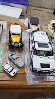Photo showing various die cast model cars including a large white Hummer, vintage style 1930s Ford models in yellow and cream, and smaller silver and white resin/ceramic models on a table.