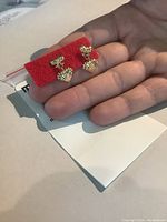 Front view of the 14k yellow gold earrings showing bow posts with heart-shaped drop pendants held by hand.