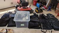 Overview of sweaters, pants, and plastic bin containing additional clothing items laid out on table in shop space with lawn equipment in background