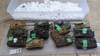 Overview of 12 military model tanks on display with white packing peanuts and a plastic tote.