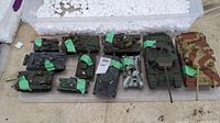 Overview of 11 military tank models displayed in a clear plastic tote, several with green tape markings and name plates, some showing loose or missing parts.