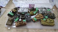 Overview of 13 different military vehicle models placed in a clear plastic tote. Variety of tank colors including green, tan, gray, and white UN vehicle.