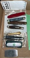 Photo showing seven pocket knives in a cardboard box, some with branding labels visible such as Old Timer and advertising blades.