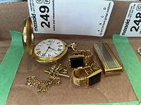 Overall view of lot including open Dynasty pocket watch, gold-tone money clip, and cuff links with black stones, all on cardboard base with auction label.