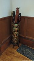 Full view of tall ornate umbrella stand with assorted canes inside, placed in corner beside wood paneling, includes rubber shoe mat on floor.
