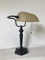 Front side view of antique style banker’s lamp showing bronze finish metal base and amber glass shade with pull chain.